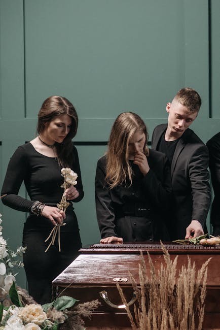 Three individuals are gathered around a wooden casket at a funeral service, with the setting illuminated by soft, natural lighting. The woman on the left holds a small floral arrangement of white flow