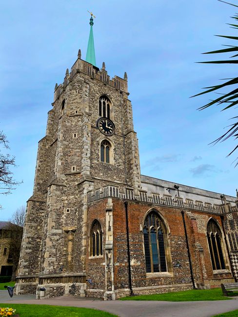 A tall, historic church tower with a pointed teal spire, constructed from a mix of grey stone and red brick, features large arched stained glass windows and a clock face displaying the time as 12:05. 
