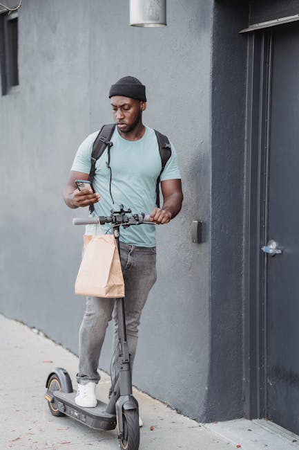A young man with dark skin, wearing a black beanie, a light blue T-shirt, and grey jeans, is standing outdoors against a grey wall. He is holding a smartphone in one hand and a brown paper bag in the