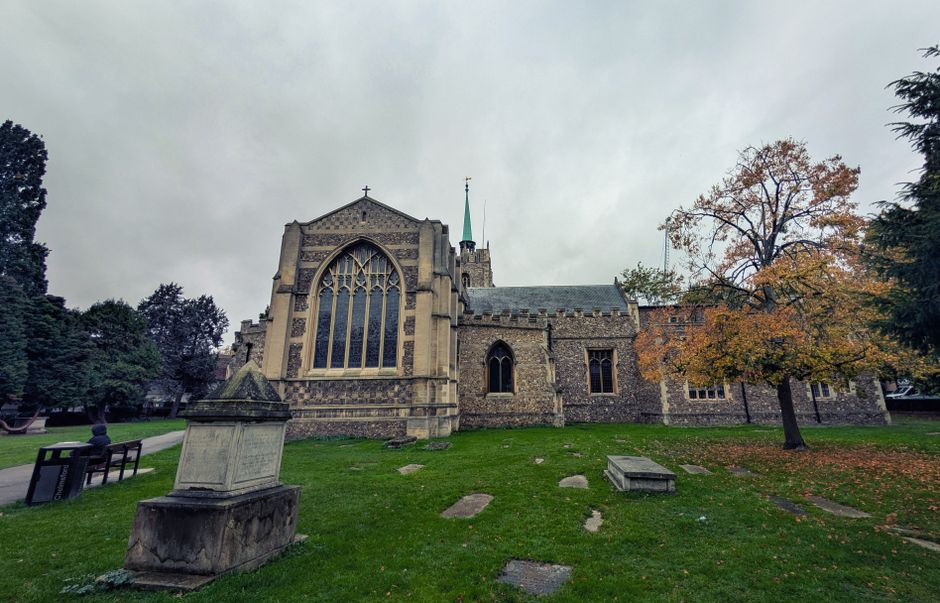 A large stone church with Gothic architectural features, including tall pointed windows with intricate tracery and a steep roof. The church is set on a grassy area with scattered stone slabs and a par