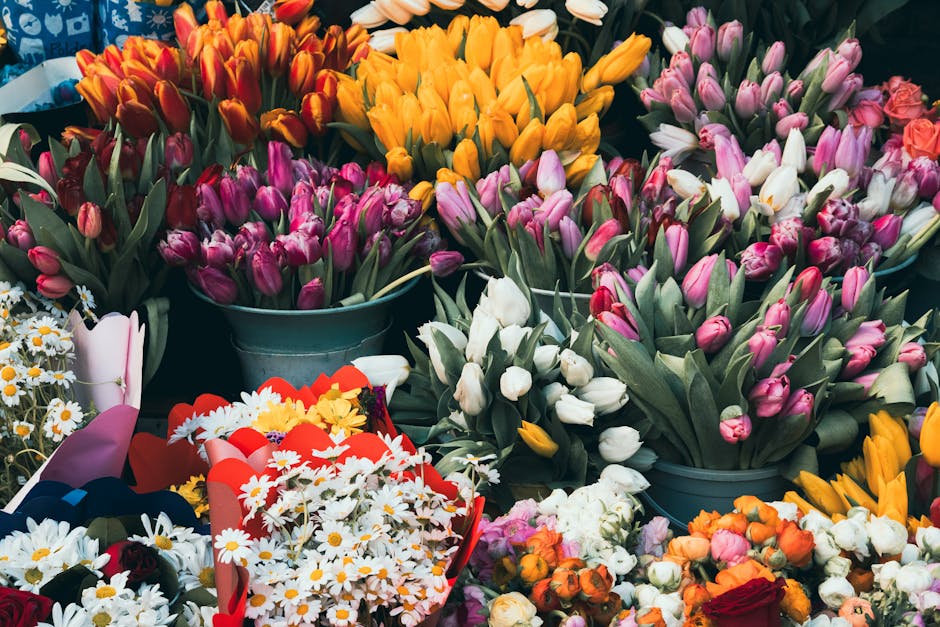 An array of colorful fresh tulips in shades of pink, white, yellow, orange, and red arranged in teal buckets and displayed on a surface. Among the tulips, there are small bouquets of daisies with whit