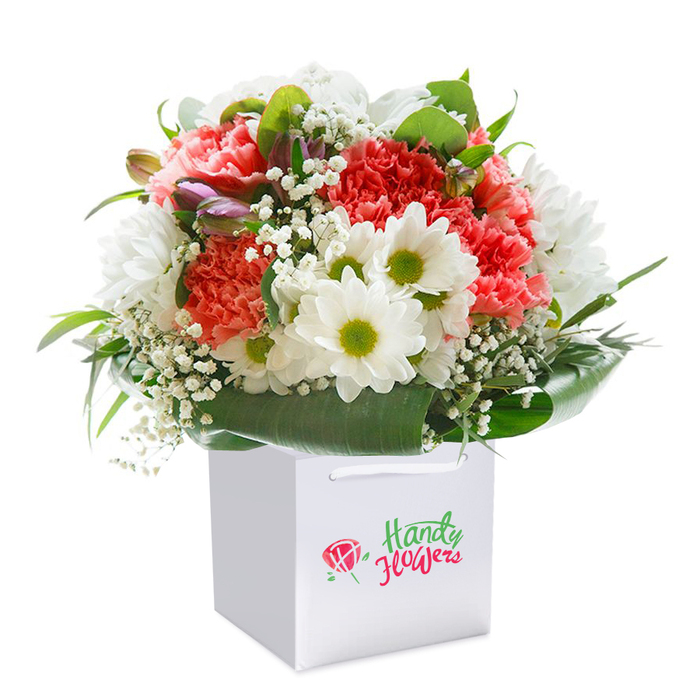 An expertly crafted bouquet, presented upright in a pristine white square gift bag, serves as a window into the artistry of Chelmsford florists. This arrangement weaves together the exuberance of soft coral and peach carnations-each petal layered and ruffled for maximum texture-with the crispness of white daisy chrysanthemums whose lime-green centers seem to glow with vitality, much like blooms you'd see brightening the stalls along High Street or the riverside paths. Threaded elegantly throughout are airy trails of pure white gypsophila, lending a delicate, cloud-like softness and a sense of weightlessness to the composition. A sprinkling of small, unopened purple-tinged buds peeks from the side, introducing layers of intrigue and dimensionality to the arrangement. Enveloping the flowers, broad glossy leaves in deep greens form a natural, protective collar that mirrors the lush landscapes of Central and Beaulieu Park. Held securely by smooth white ribbon handles, the bouquet's presentation is both thoughtful and practical-ready for hand delivery to offices along Parkway or homes nestled in Moulsham Lodge. The branding on the bag is seamlessly integrated, not distracting from the artistry of the floral display itself. Set against a flawless white background that makes each colour and petal pop, this bouquet radiates freshness, care, and a touch of luxury, ideal for moments that matter-be it a Chelmsford birthday, anniversary, or spontaneous gesture of appreciation.
