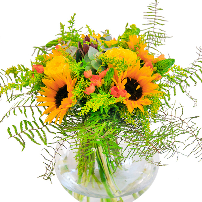 The first impression of this sun-drenched arrangement is exuberant cheerfulness-a celebration of colour and life crafted with the experienced hands of a Chelmsford florist. Nun's sunflowers command attention, their dark centers framed by layers of vivid yellow-orange petals that nearly glow beneath the steady light, reminiscent of a stroll through Hylands Estate at peak bloom. Intertwined are sumptuous yellow roses, their tightly whorled petals forming rosette shapes that add structure and classical beauty against the everchanging green. Touches of bright, rounded red hypericum berries gleam among the foliage, each glistening orb contrasting the soft textures of the blossoms. Airy sprigs of pale green solidago, feathery ferns, and slender eucalyptus leaves tumble out along the base, imbuing the whole with a freshness akin to the gardens bordering Central Park. Encased in a pristine spherical glass vase, the stems create a verdant lattice, visible through clear water and evoking a sense of grounded clarity. As the local light falls gently across the table, every detail is accentuated: the downy sunflower petals, the velvety curve of a rose, the crisp vitality of new greens. This bouquet is designed for those cherished celebrations-be they birthdays, new beginnings, or heartfelt gestures to neighbours throughout Old Moulsham-bringing the timeless beauty of Chelmsford's floral heritage into every home.